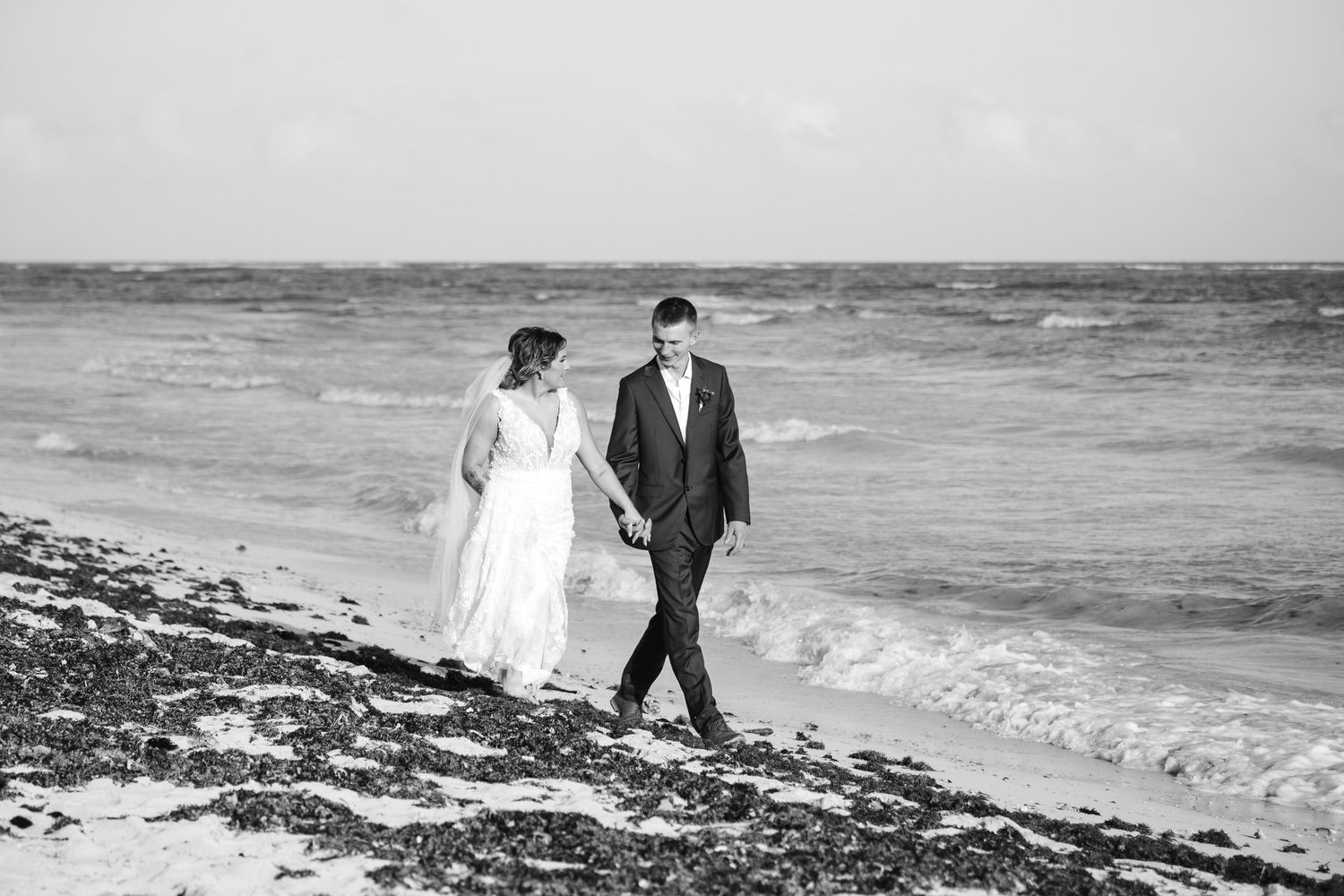 Black and white photo of bride and groom holding hands walking along ocean shoreline.