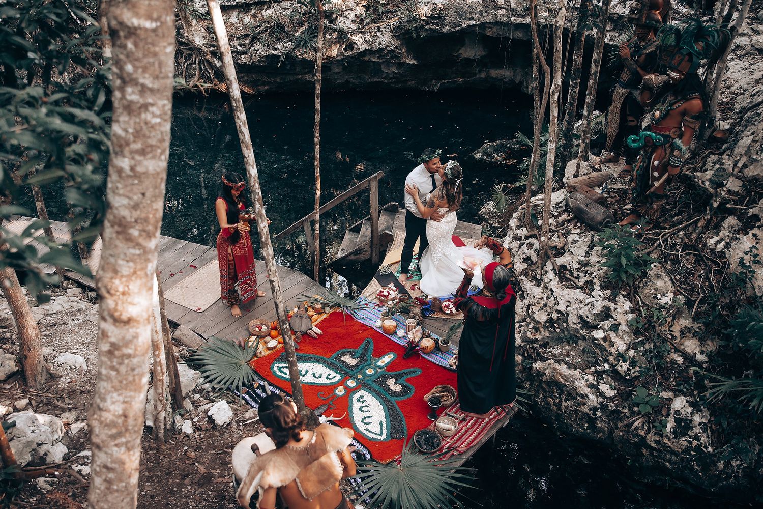 An intimate outdoor wedding ceremony in a wooded cave setting with a red carpet and guests gathered around.