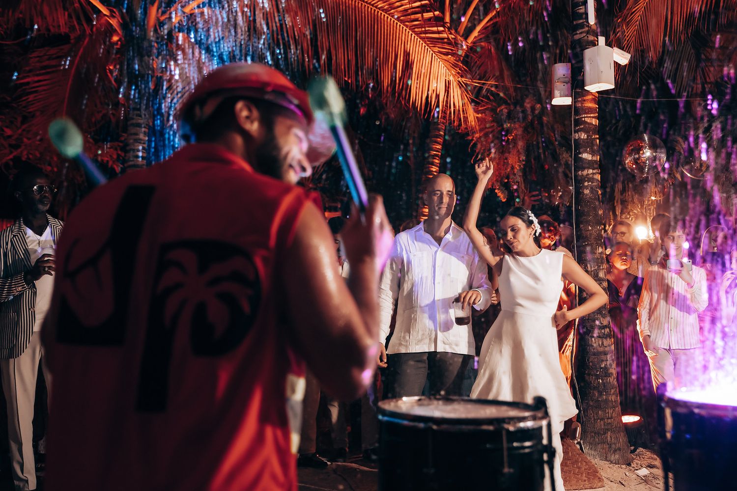 Festive dance party with guests in white attire under vibrant purple and red lighting.