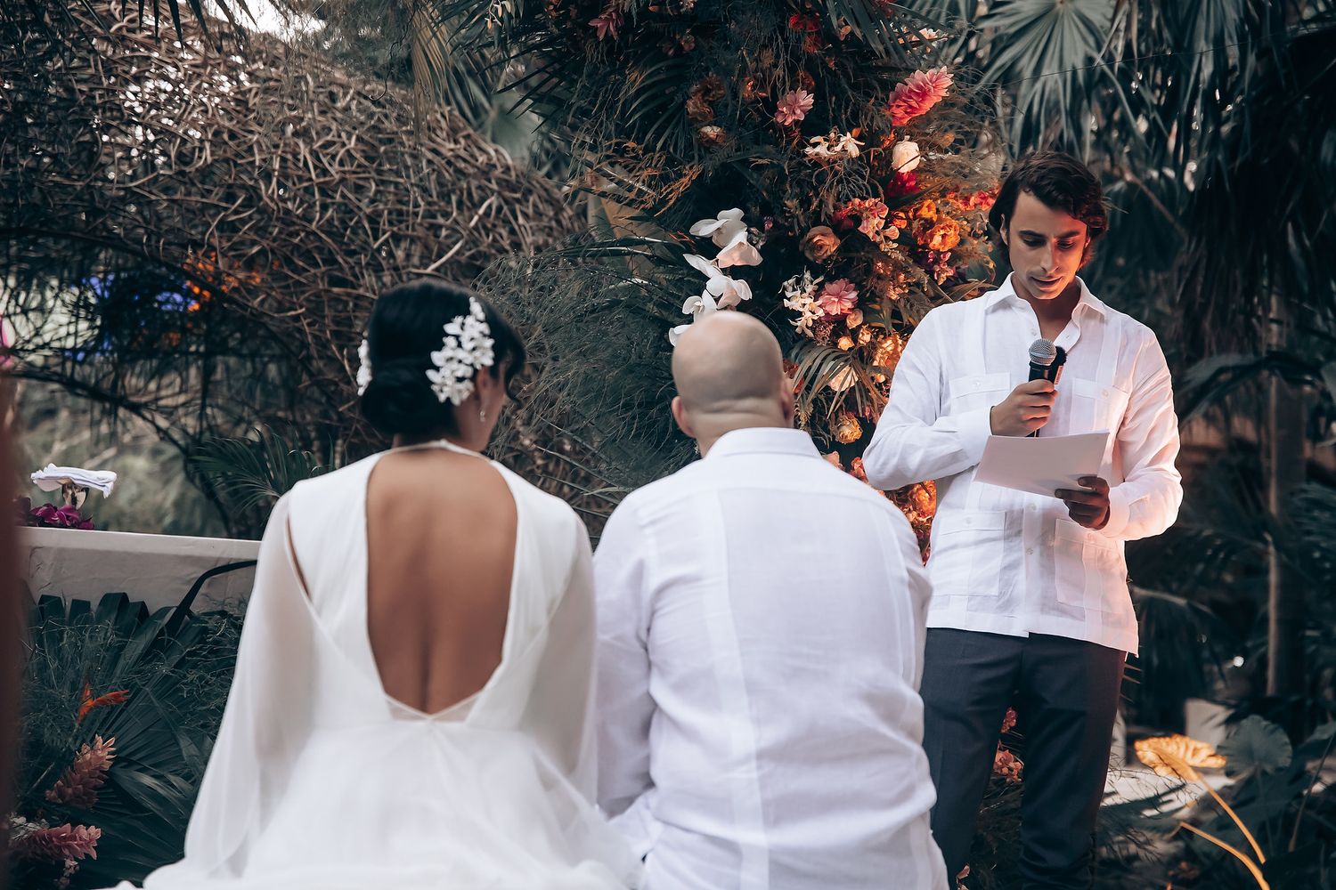 Couple and officiant stand together during outdoor wedding ceremony with floral decorations.