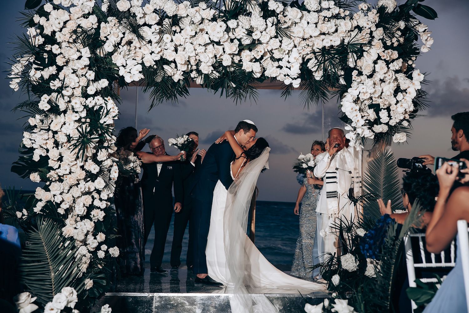 Wedding ceremony under a white floral arch with ocean backdrop at sunset.
