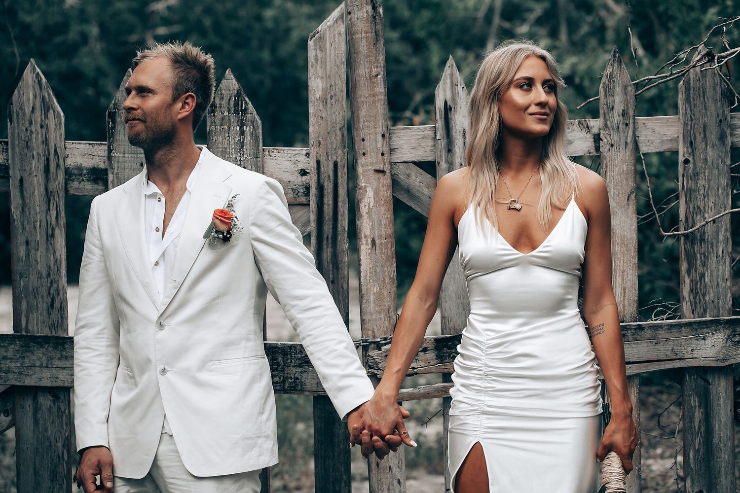A stylish couple in white attire poses against a weathered wooden fence.