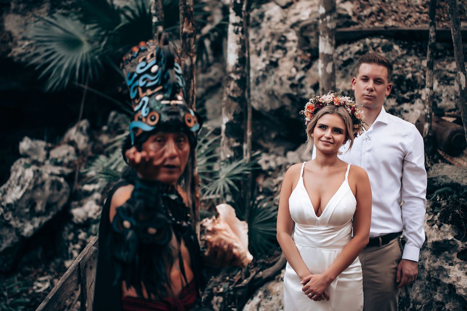 A wedding couple poses against a stone wall backdrop while a photographer captures the moment in a tropical setting.