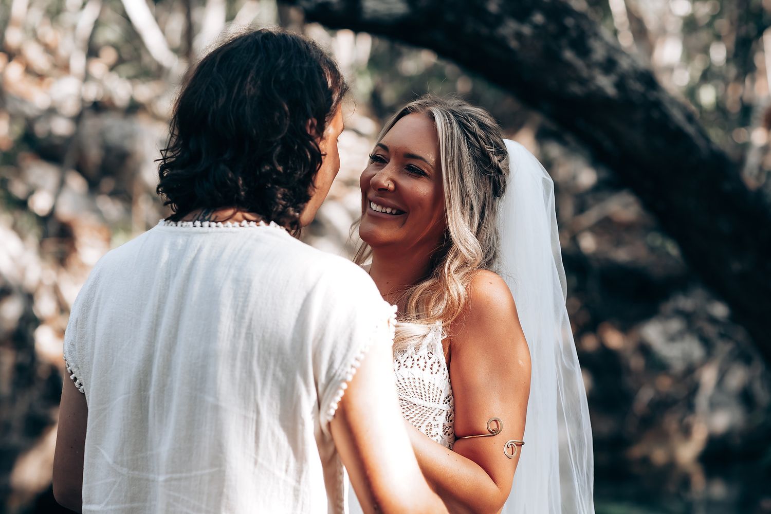 A couple in wedding attire share an intimate moment under trees with sunlight filtering through the branches.