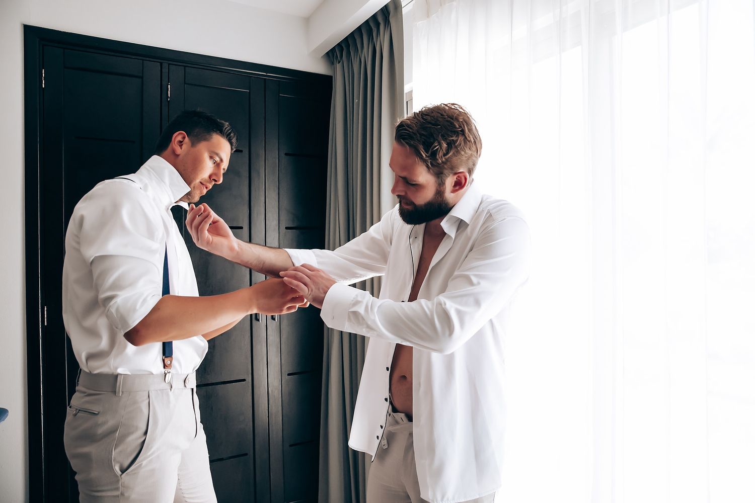A person in a white shirt helps another person adjust their sleeves in a room with dark doors and white curtains.