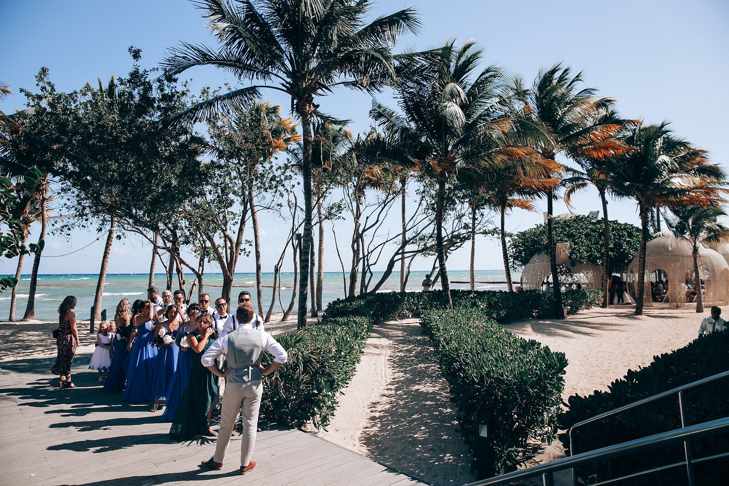 Palm trees line a tropical beach walkway with white sand and turquoise waters in the distance.