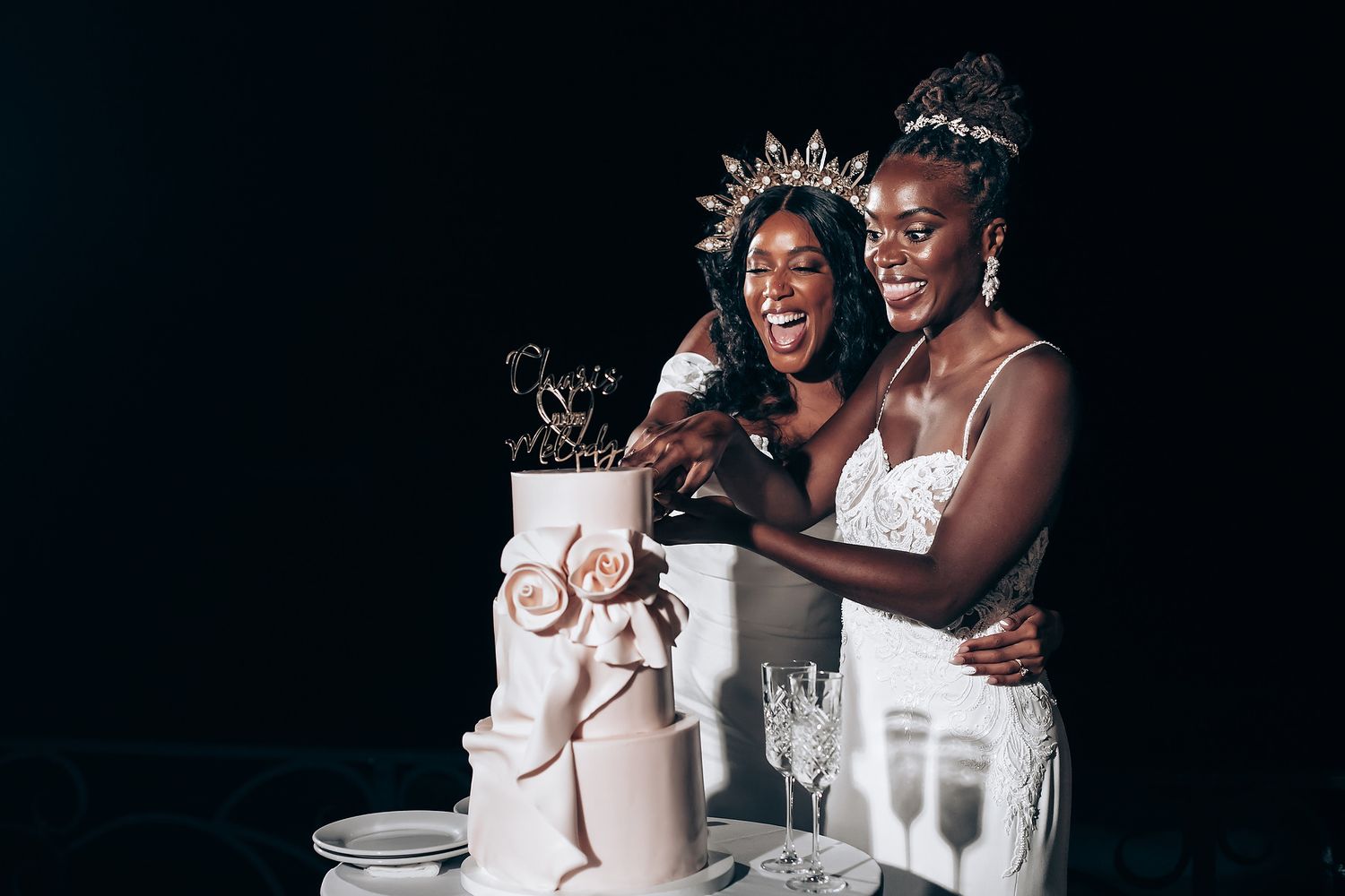 Three women in white dresses share laughter and embrace during a celebration against a dark background.