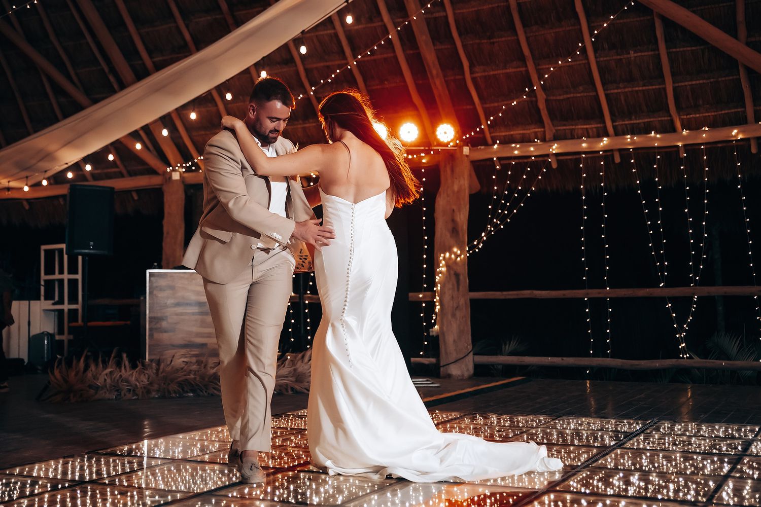 Wedding couple embraces on dance floor under romantic string light canopy in rustic venue.