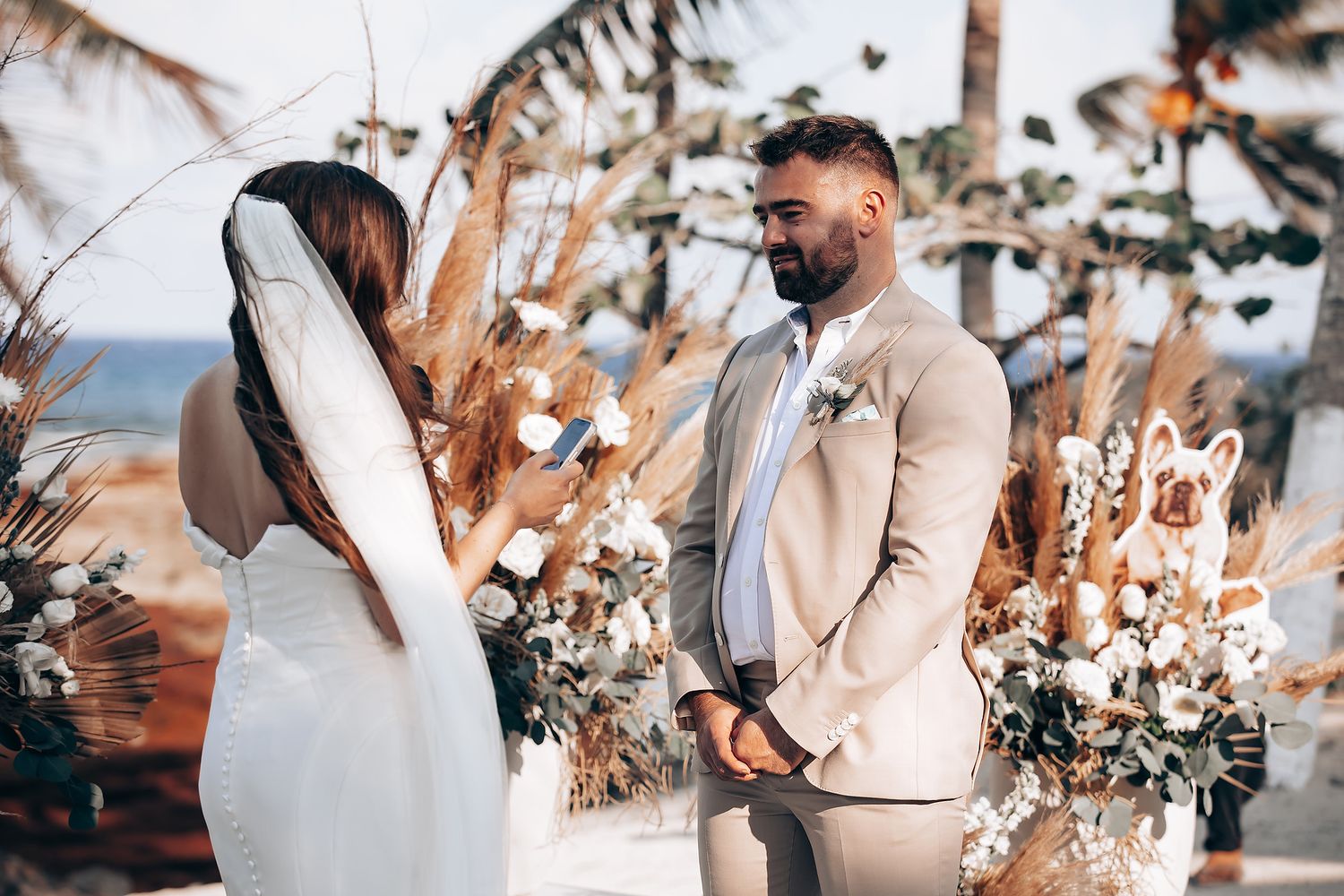 A couple exchanges vows at a tropical beach wedding ceremony decorated with dried palm leaves and pampas grass arrangements.