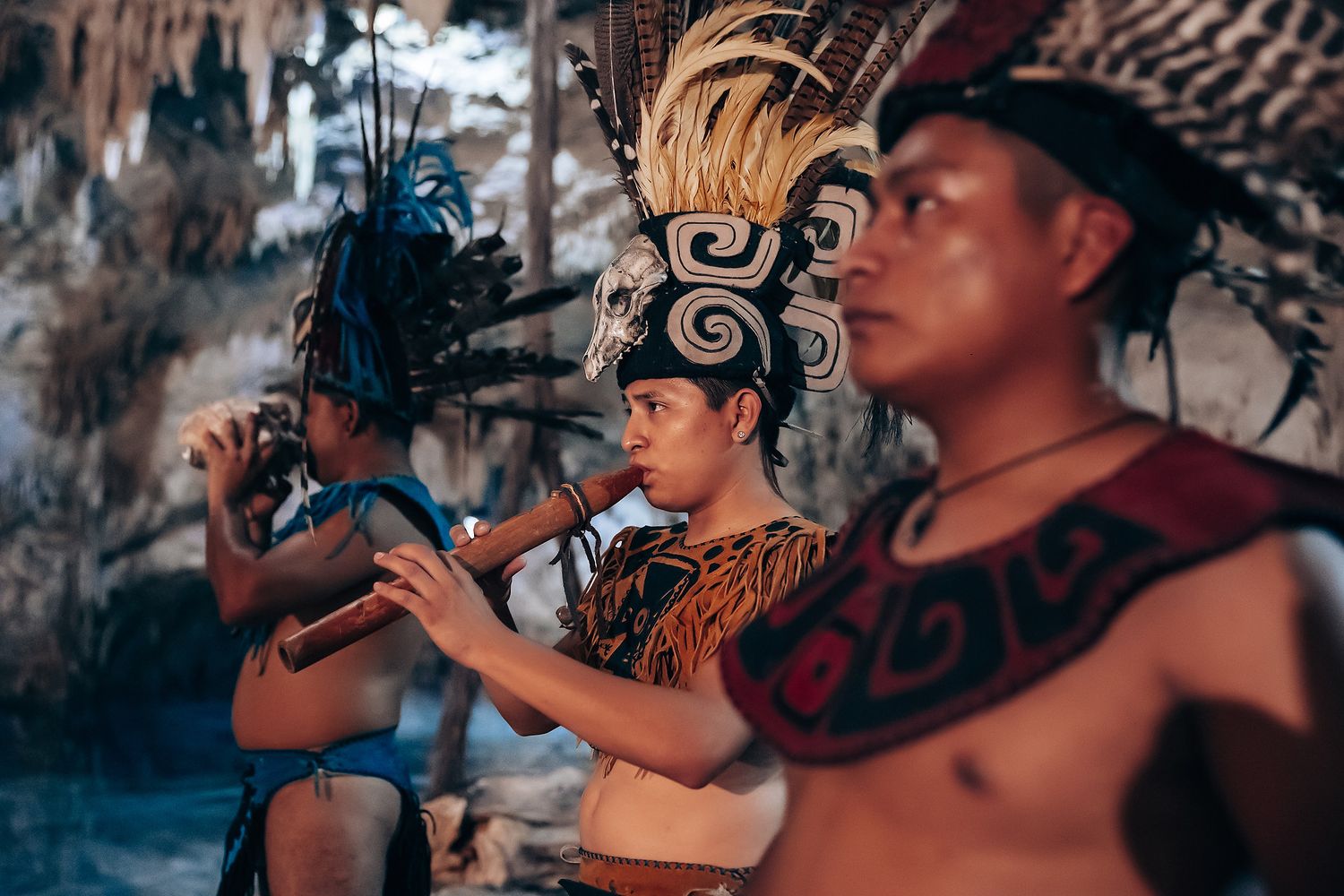 A person wearing a traditional feathered headdress performs with a musical instrument in a ceremonial setting.