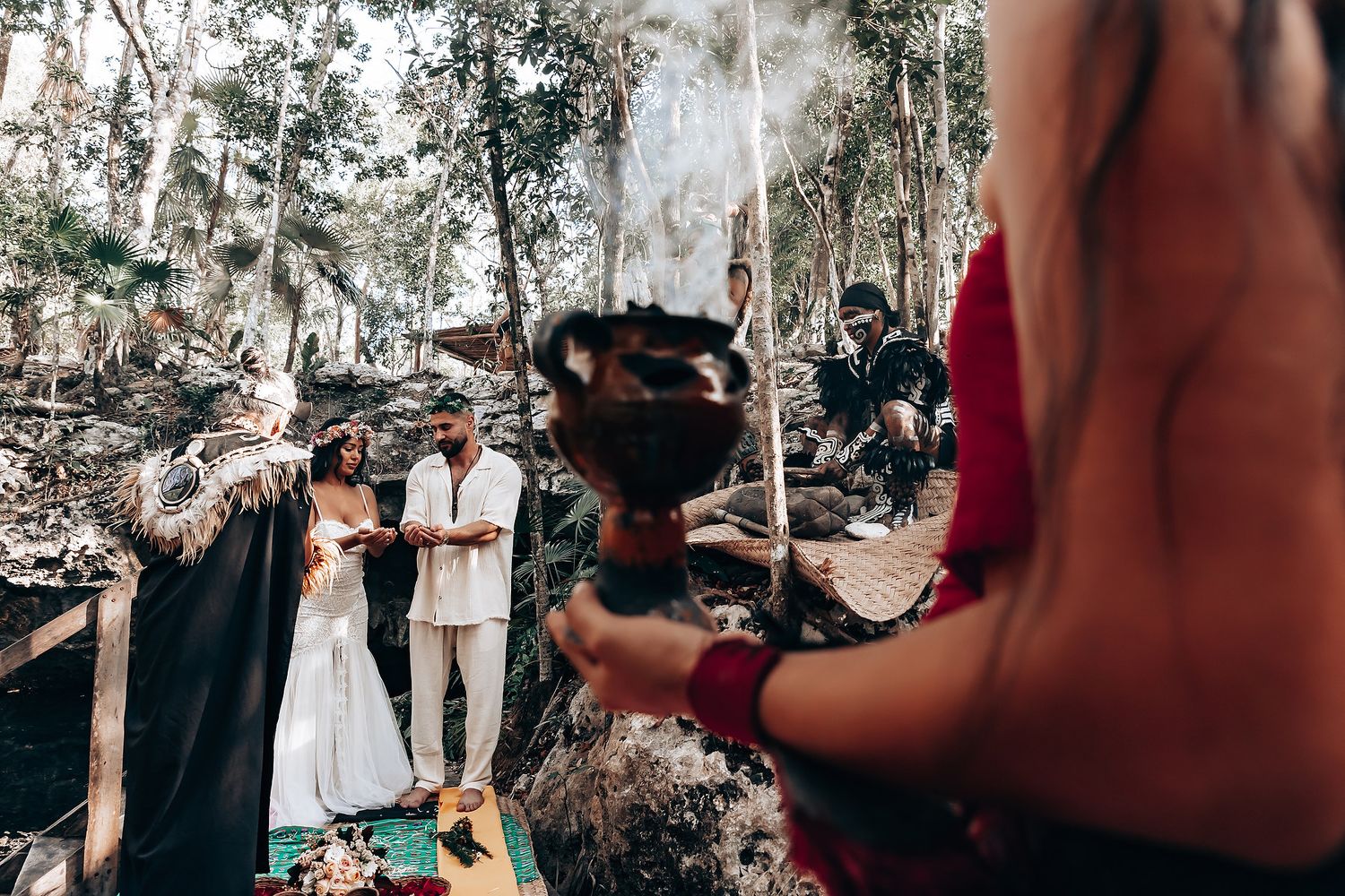 A wedding ceremony takes place in a wooded outdoor setting with smoke and natural decor.
