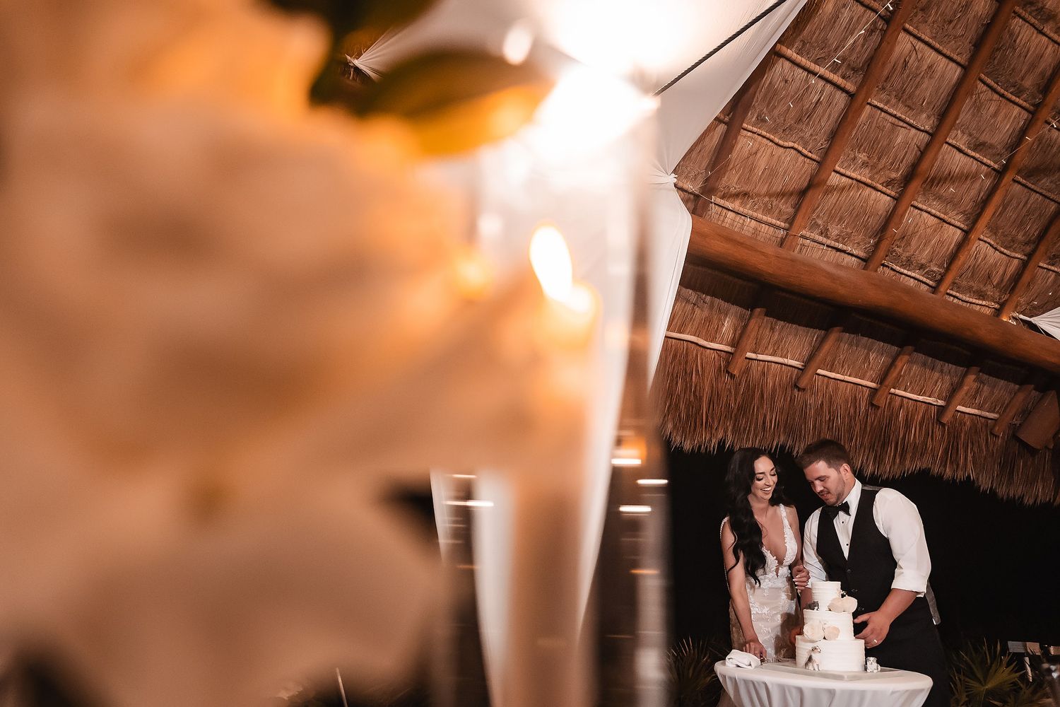 Bride and groom cutting wedding cake under thatched palapa roof at tropical outdoor reception venue.