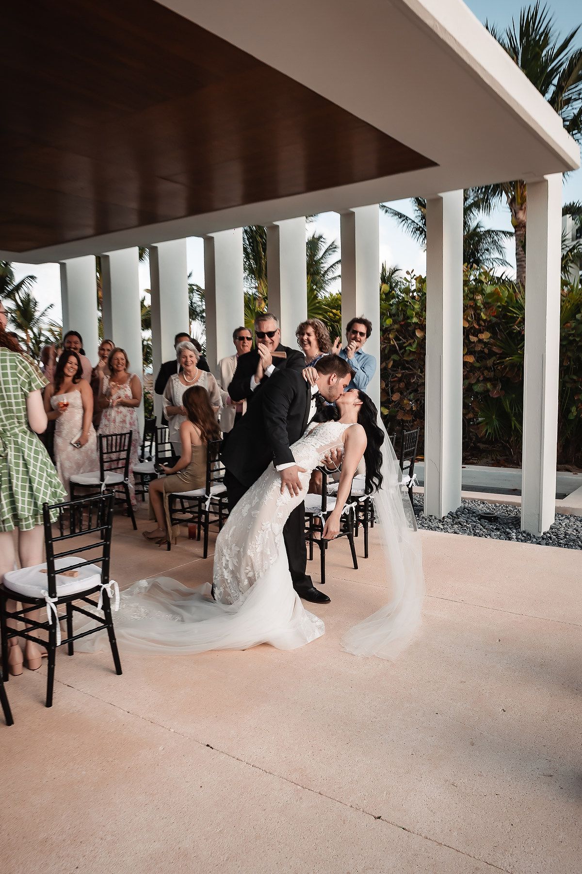 Groom dips bride for a kiss at outdoor wedding ceremony with guests watching, palm trees and white columns in background.