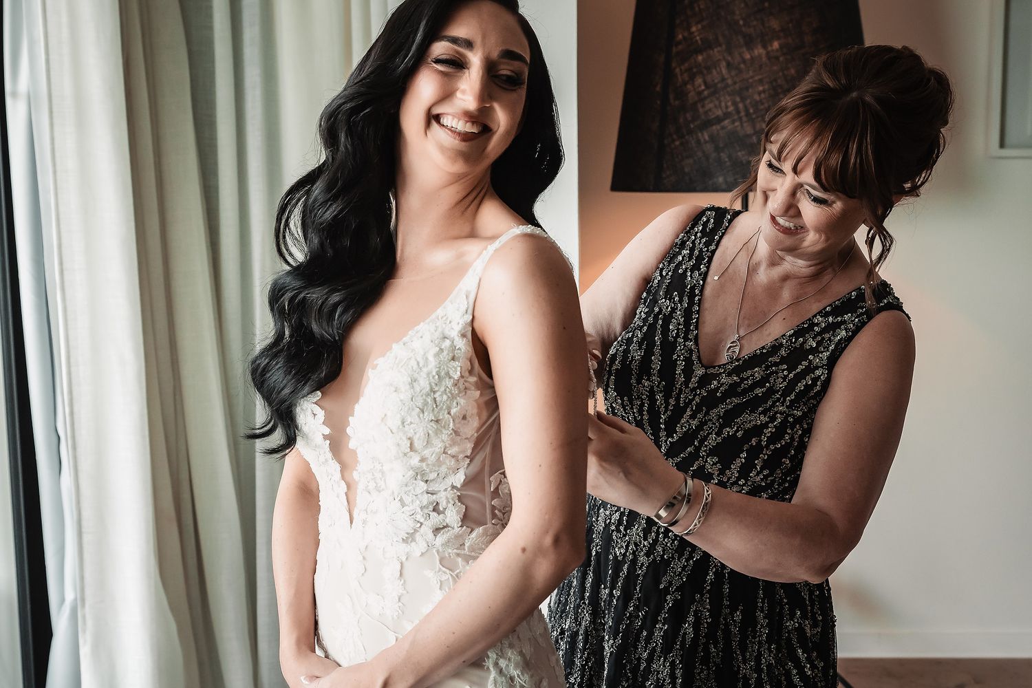 Bride in lace wedding gown smiles as older woman helps fasten her dress in a bright, elegant room.