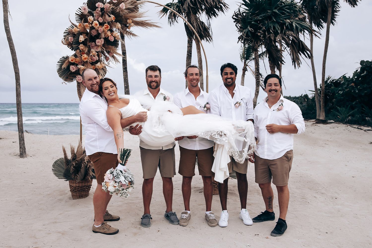 Wedding group poses for photos on a sandy beach with palm trees and ocean waves in the background.