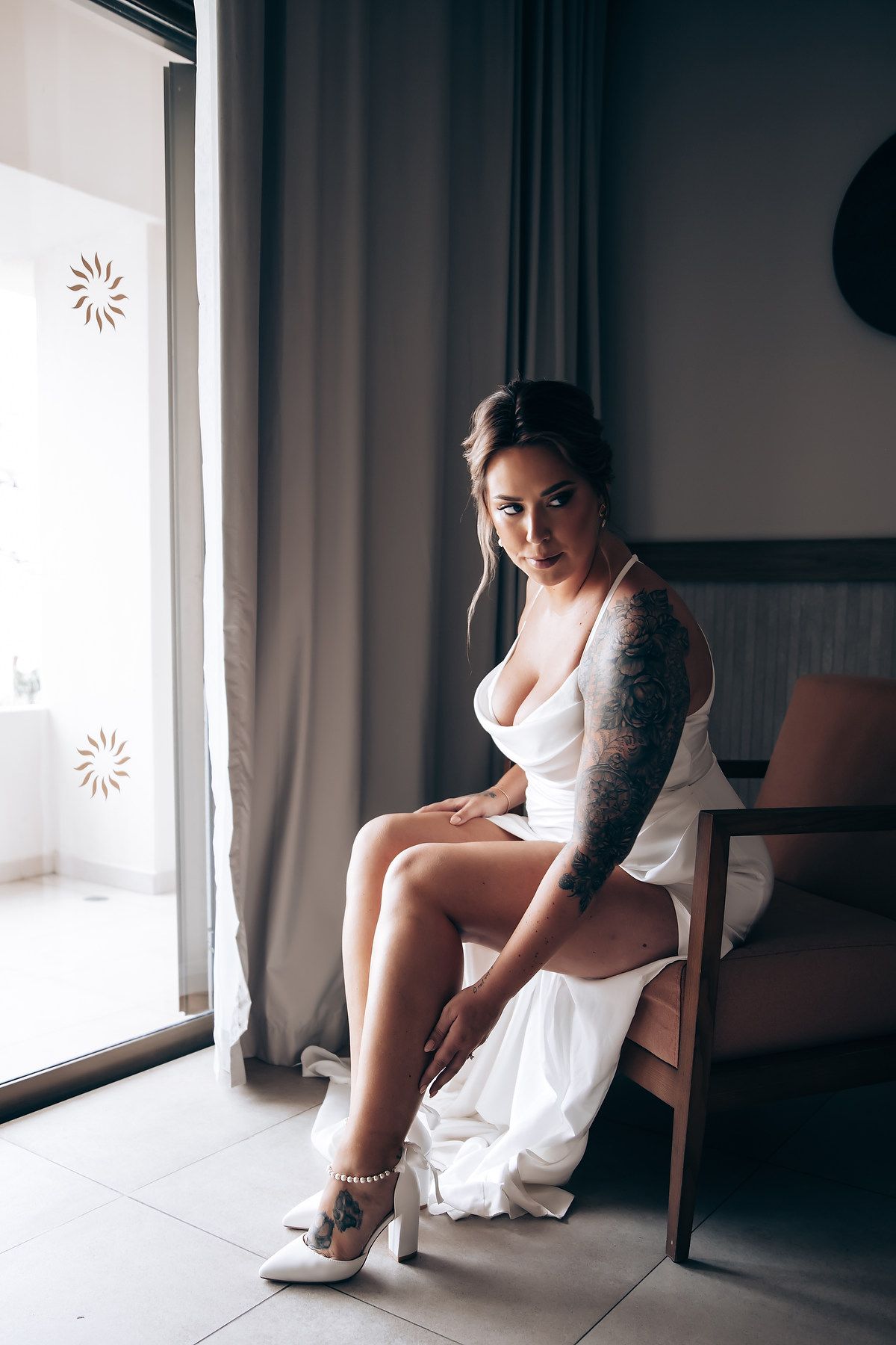 Bride in white dress sits elegantly by window during wedding day preparation.
