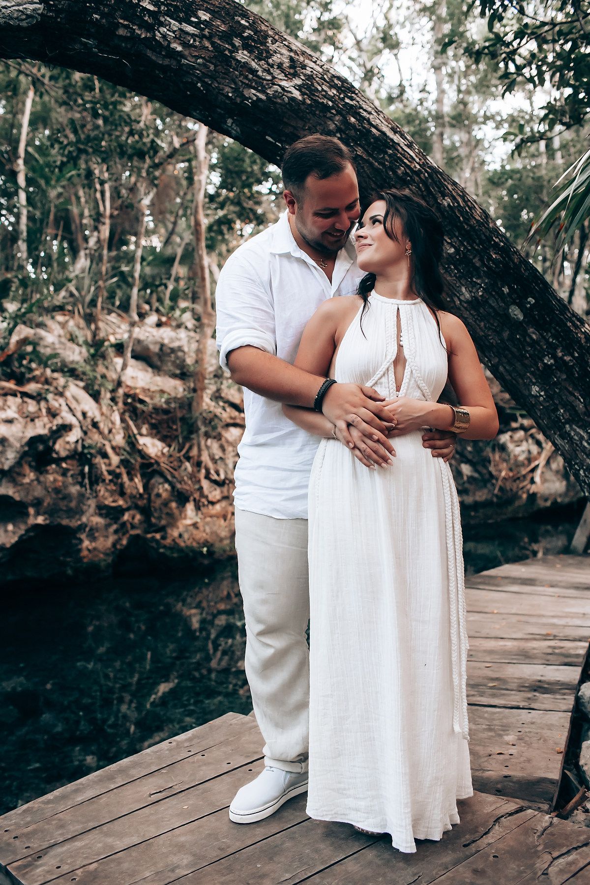 Couple in white attire embracing on wooden platform in forest setting.
