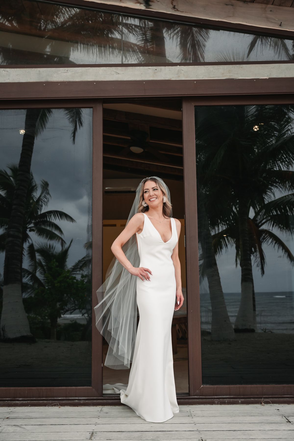 Bride in elegant white sleeveless gown and veil stands smiling in glass doorway with tropical palm trees and ocean backdrop.