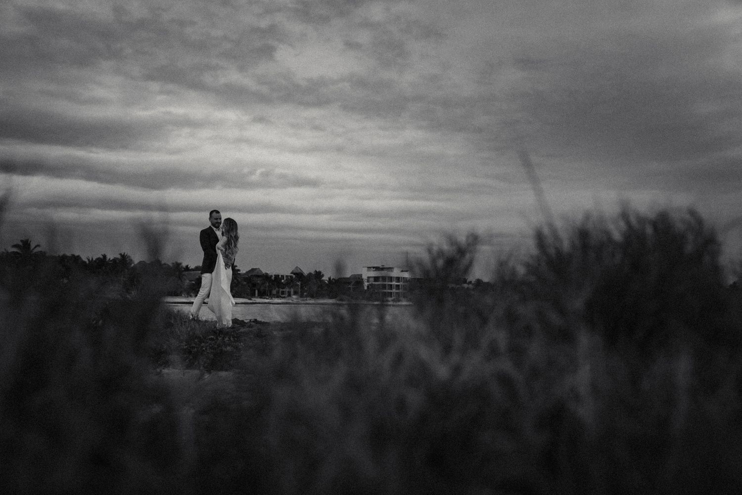 Black and white photo of bride and groom standing in a field under dramatic cloudy skies.