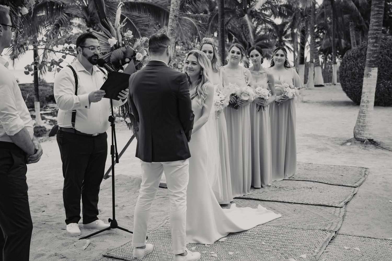 Black and white outdoor beach wedding ceremony with bride, groom, officiant, and bridesmaids.