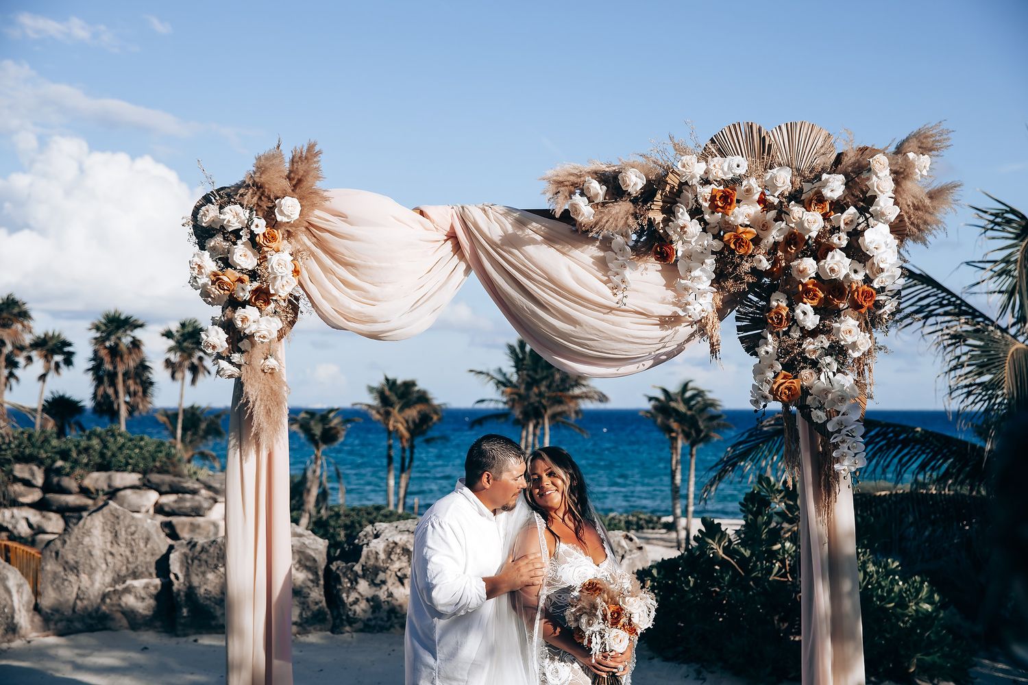 A beachside wedding ceremony setup with a draped floral arch overlooking the ocean and palm trees.