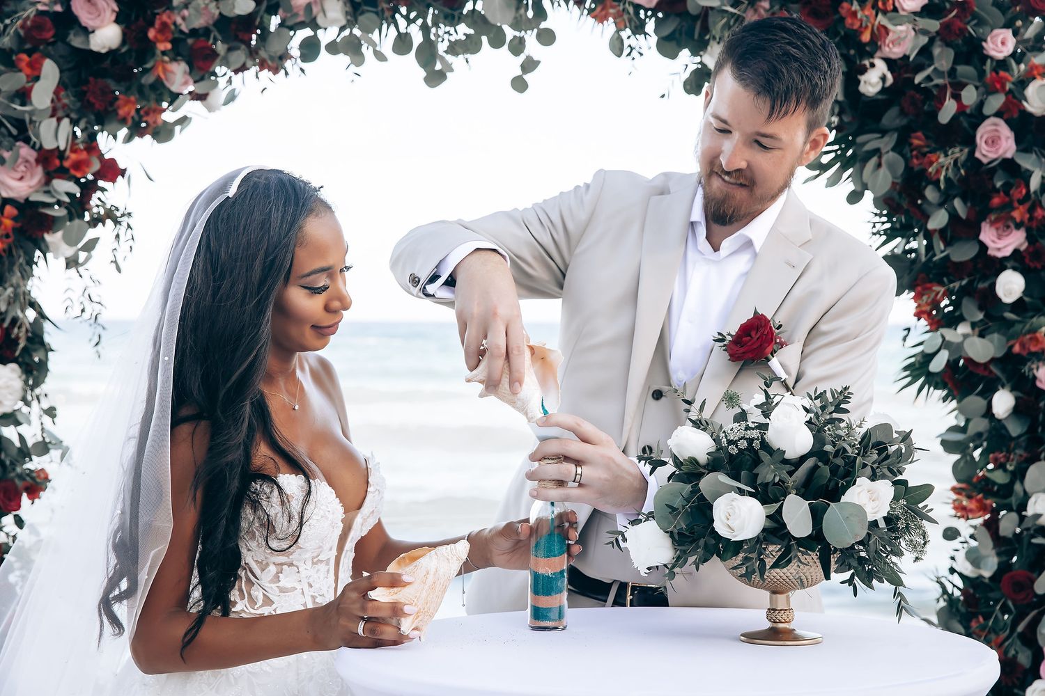 Couple shares an intimate moment under a floral arch decorated with pink roses and greenery at an outdoor wedding.