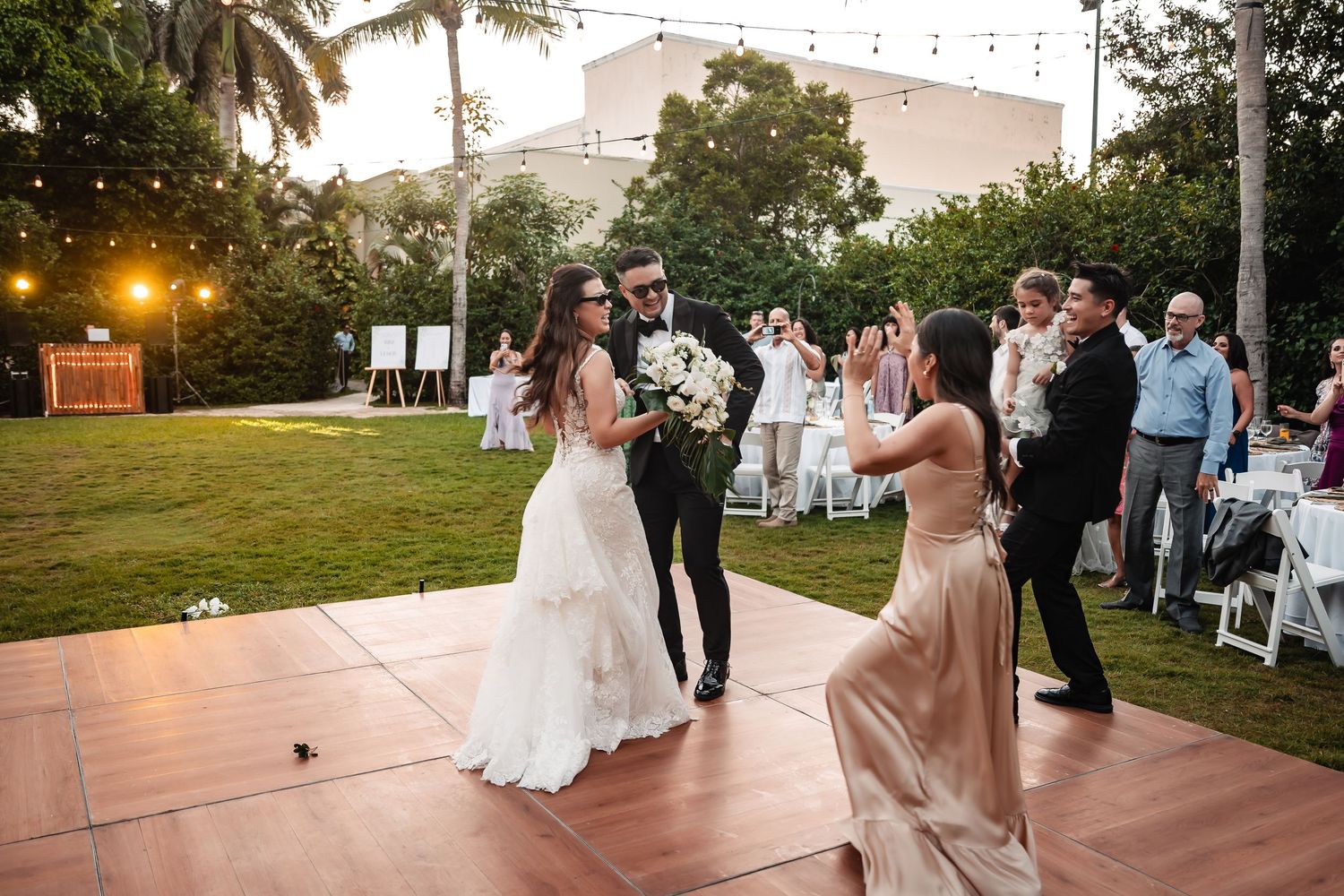 Wedding reception dance floor setup outdoors with string lights and palm trees at dusk.