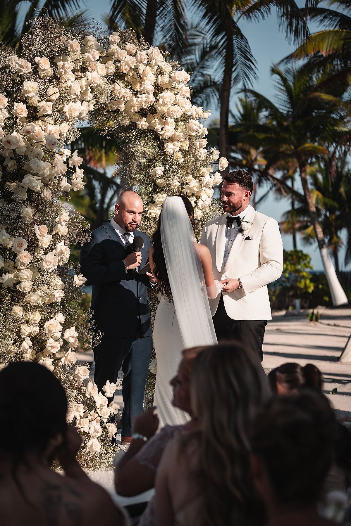 Intimate wedding ceremony moment under elaborate white floral arch with tropical beach setting.