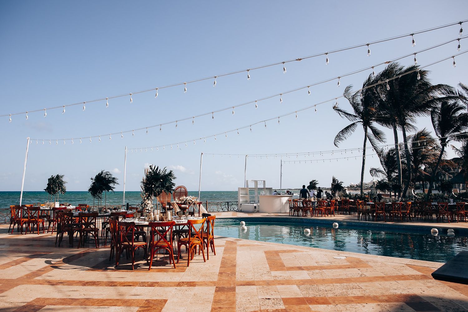 Beachfront wedding venue with string lights and wooden tables set up overlooking the ocean at sunset.