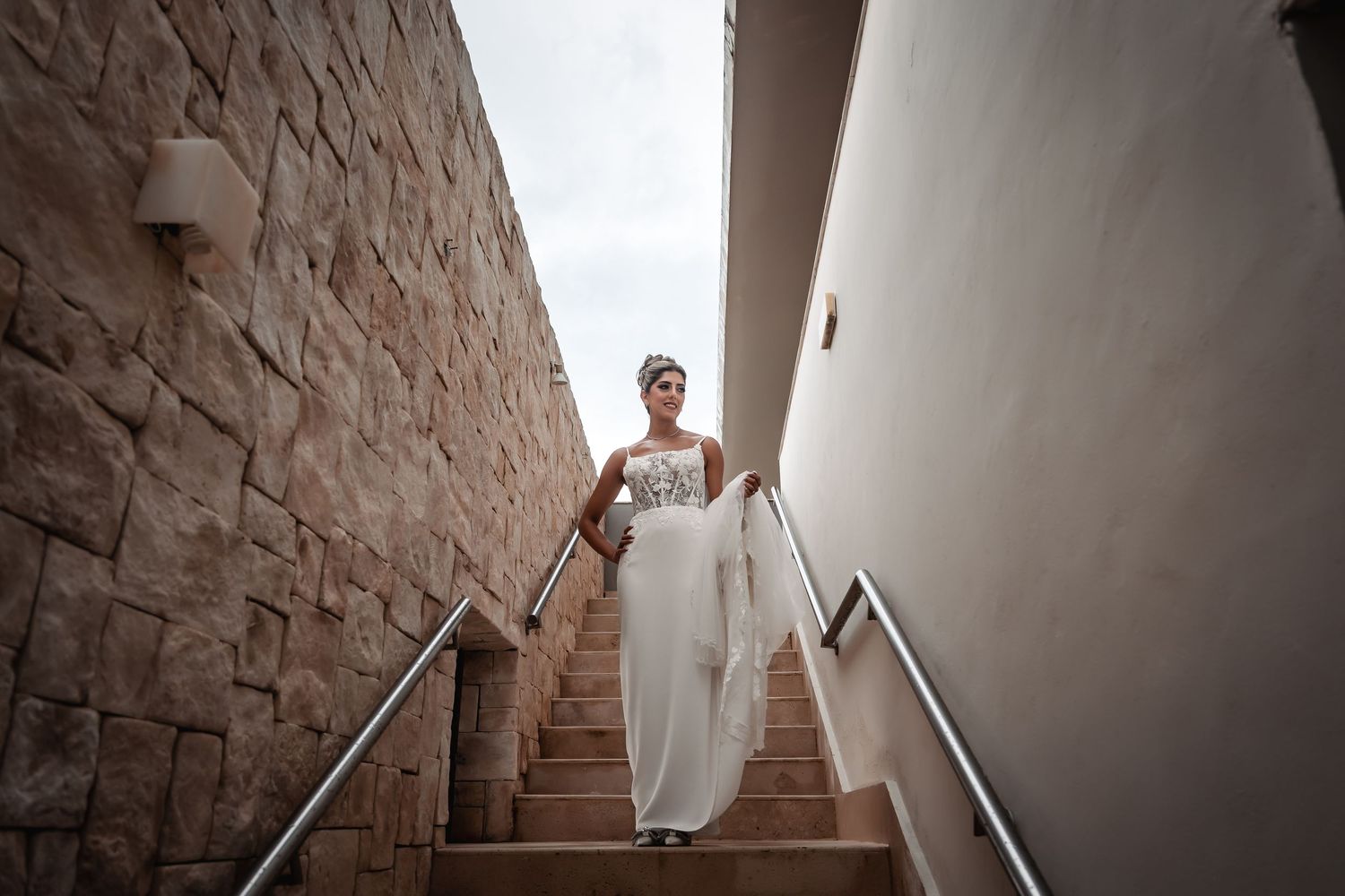 A bride in white descends a stone-walled staircase with metal handrails.