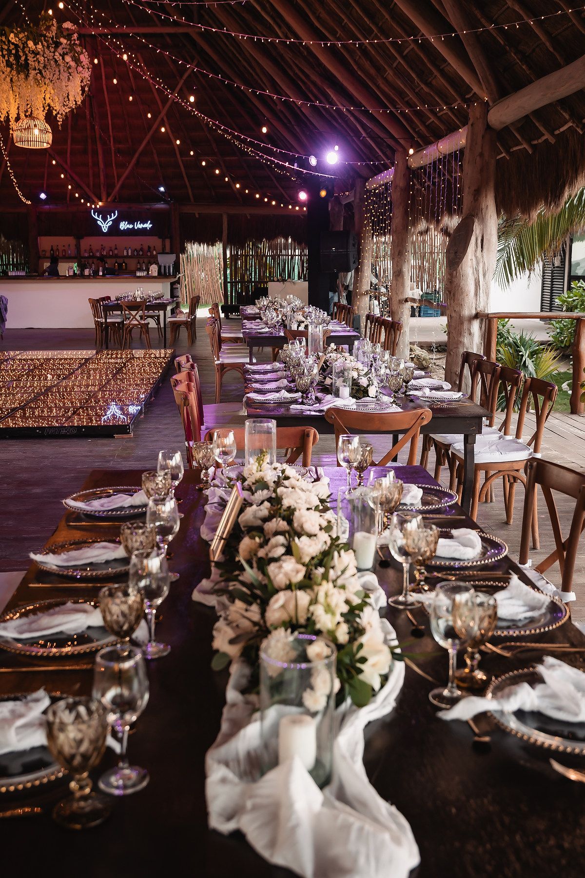 Wedding reception tables set with elegant place settings under thatched roof and string lights.