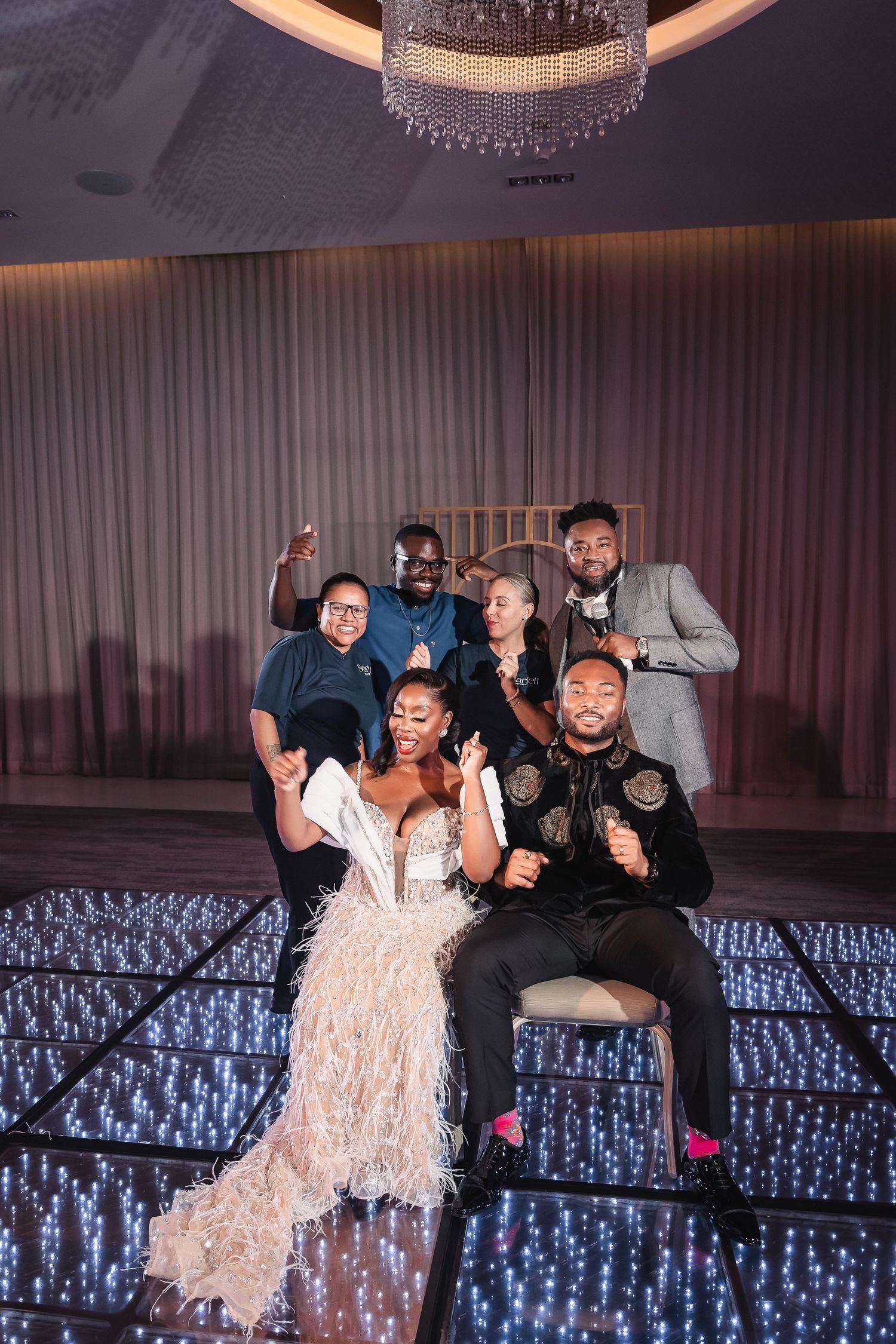 Wedding party poses playfully on illuminated dance floor under crystal chandelier.