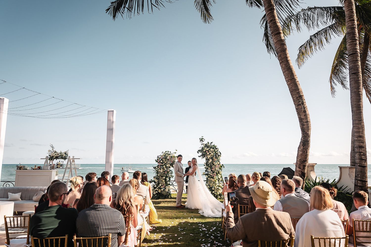 Palm trees frame an oceanfront wedding ceremony setup with rows of white chairs.