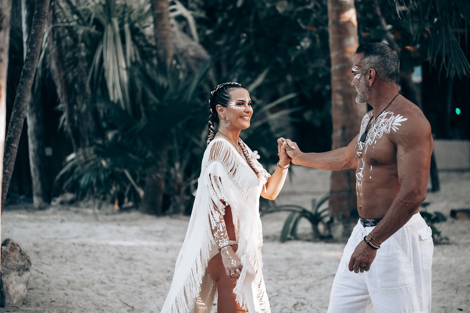 A stylish couple in white beachwear with body paint poses together on a sandy tropical path.