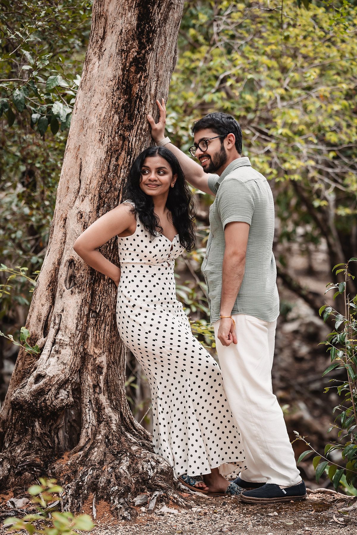 A couple poses playfully against tree trunks during an outdoor photoshoot.