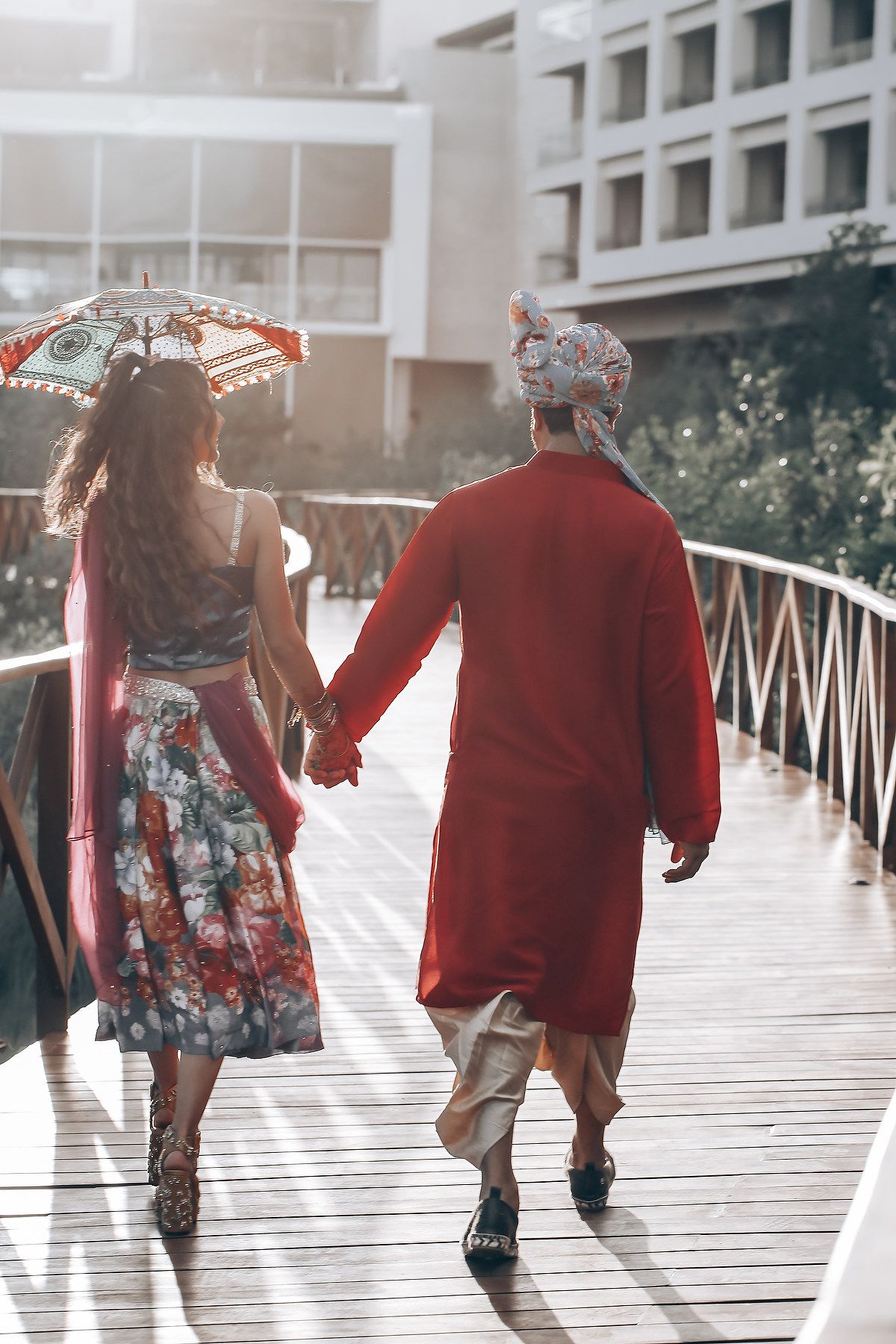 People walking along wooden boardwalk with red and floral outfits in sunlit setting.