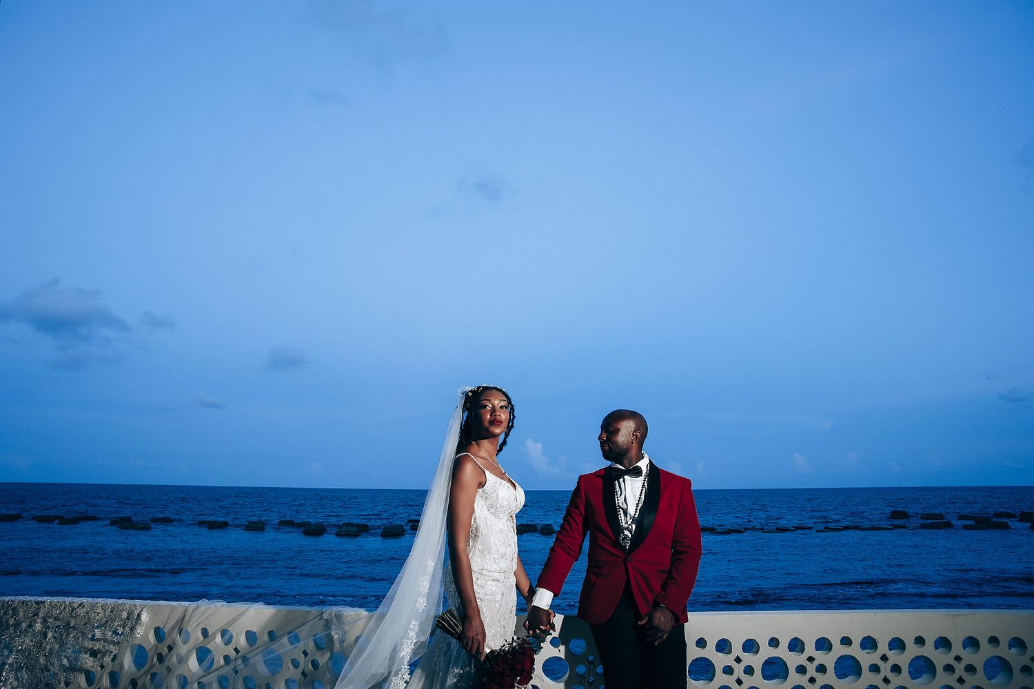 Wedding party walks along beach with ocean backdrop as bride's white dress and bridesmaids' red gowns flow in breeze.