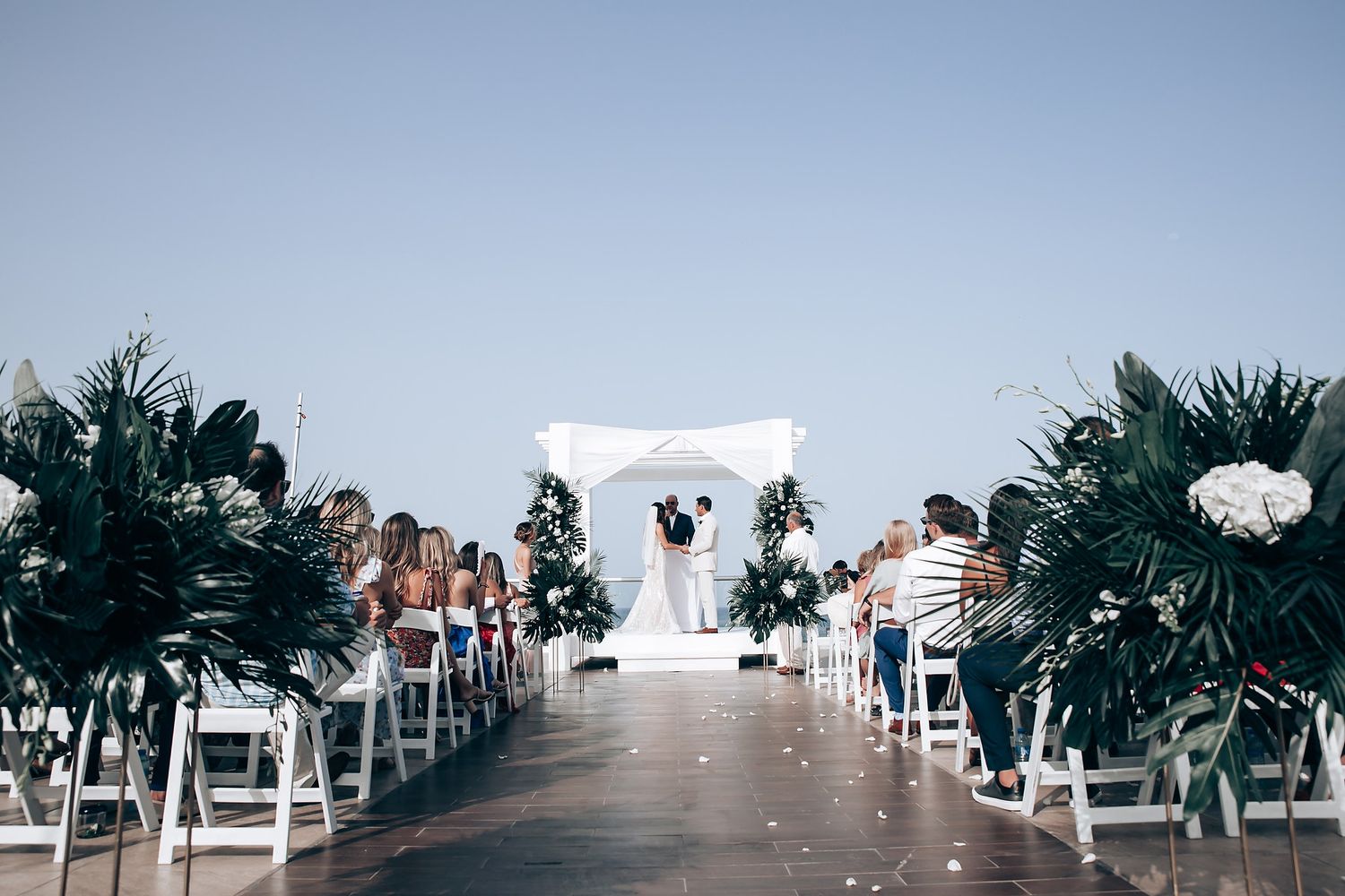 White wedding ceremony chairs arranged in rows on a beachfront with tropical palm decorations.