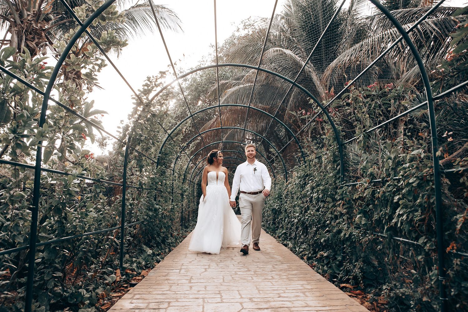 A couple walks through a lush garden archway tunnel covered in tropical greenery and vines.