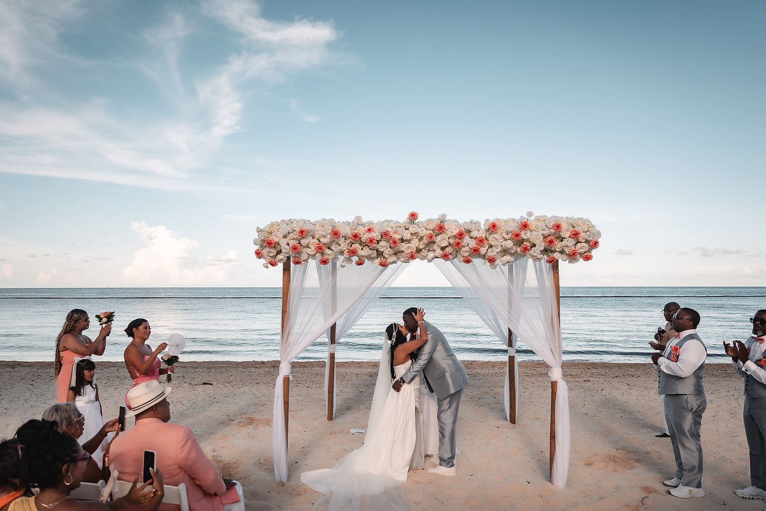 Wedding ceremony on a beach with white arch decorated in peach flowers against blue sky.