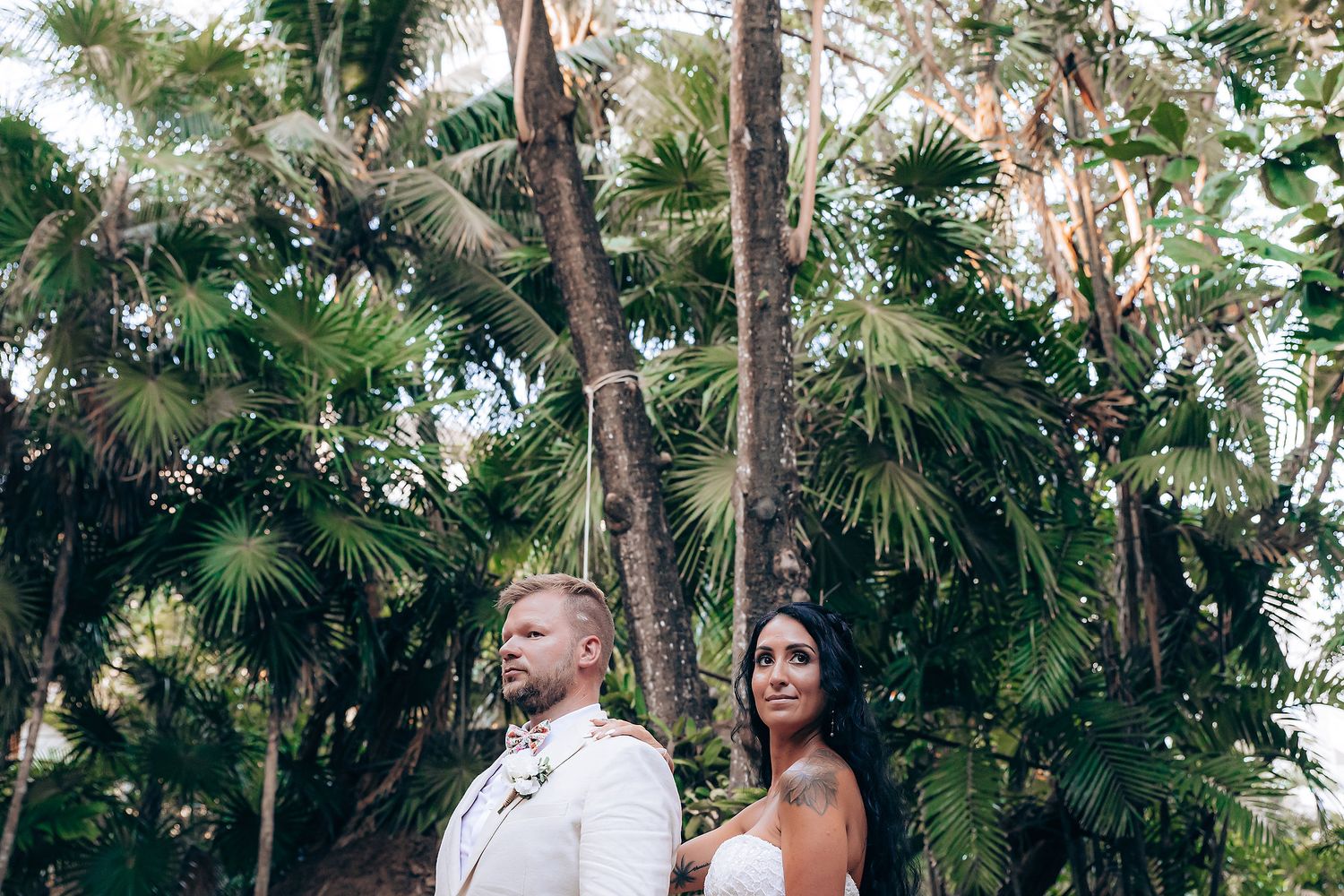 Wedding couple standing among tall palm trees in a tropical setting.