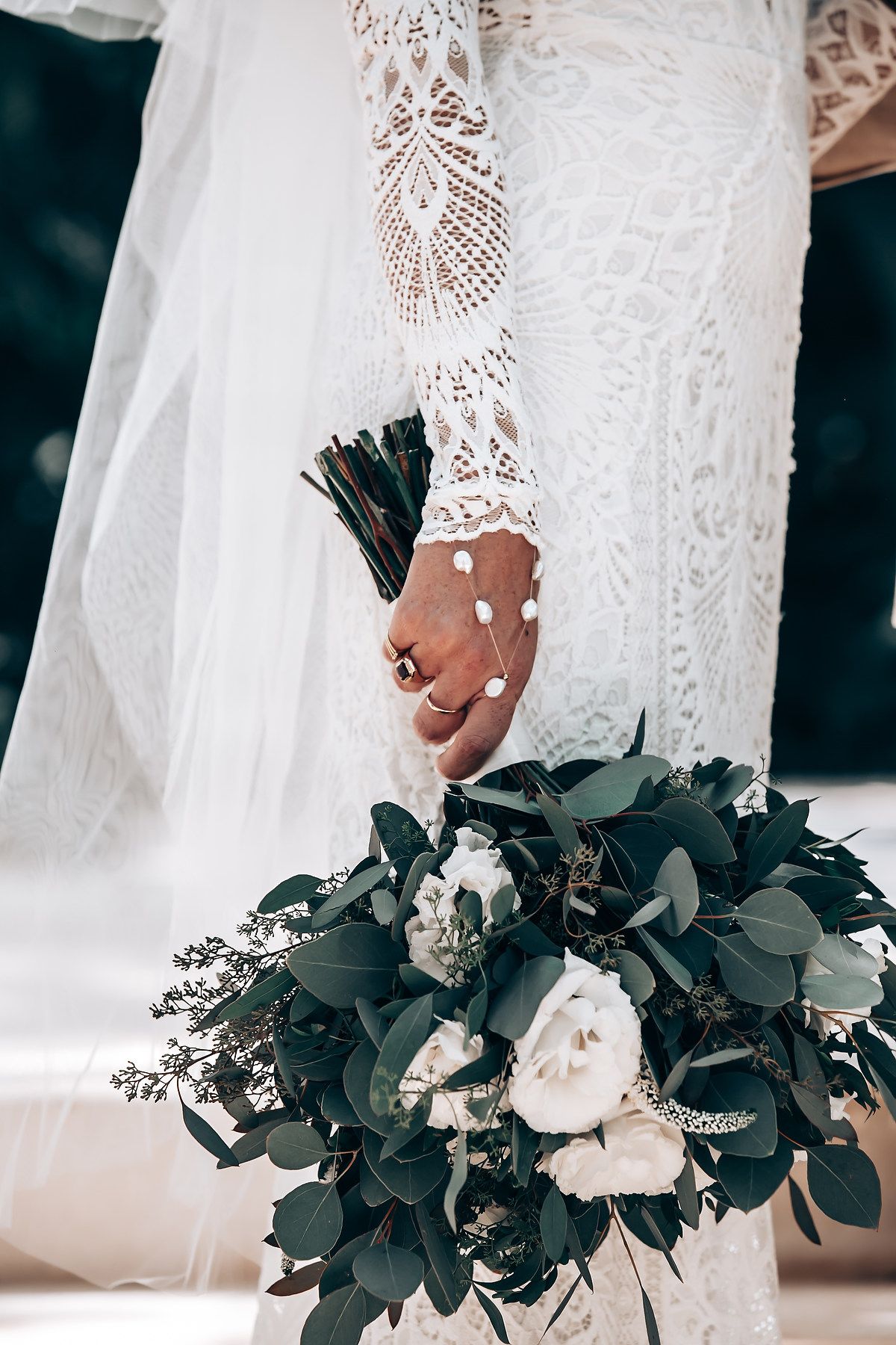 Close-up of a bridal bouquet with white flowers and dark green foliage held by hands in delicate lace sleeves.