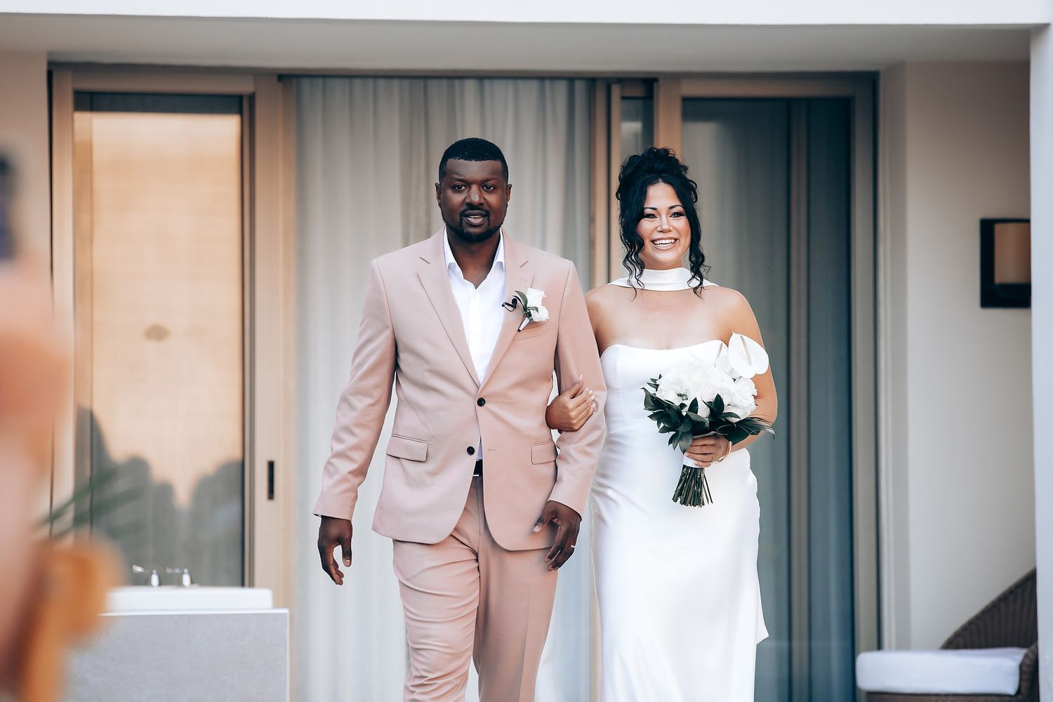 Wedding couple walks together in elegant attire of blush pink suit and white gown.