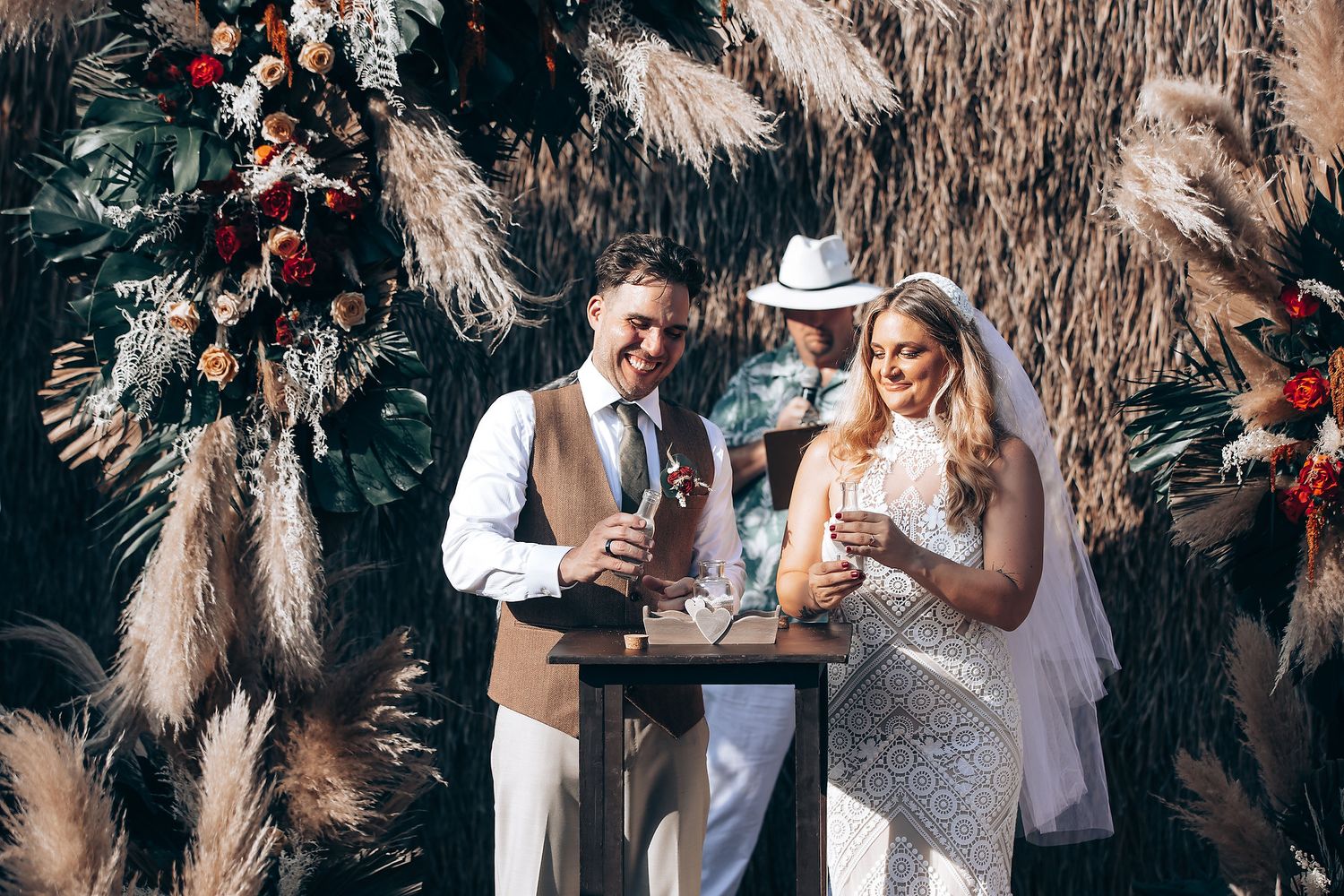 A couple in elegant white attire share drinks at a wedding ceremony decorated with pampas grass and floral arrangements.