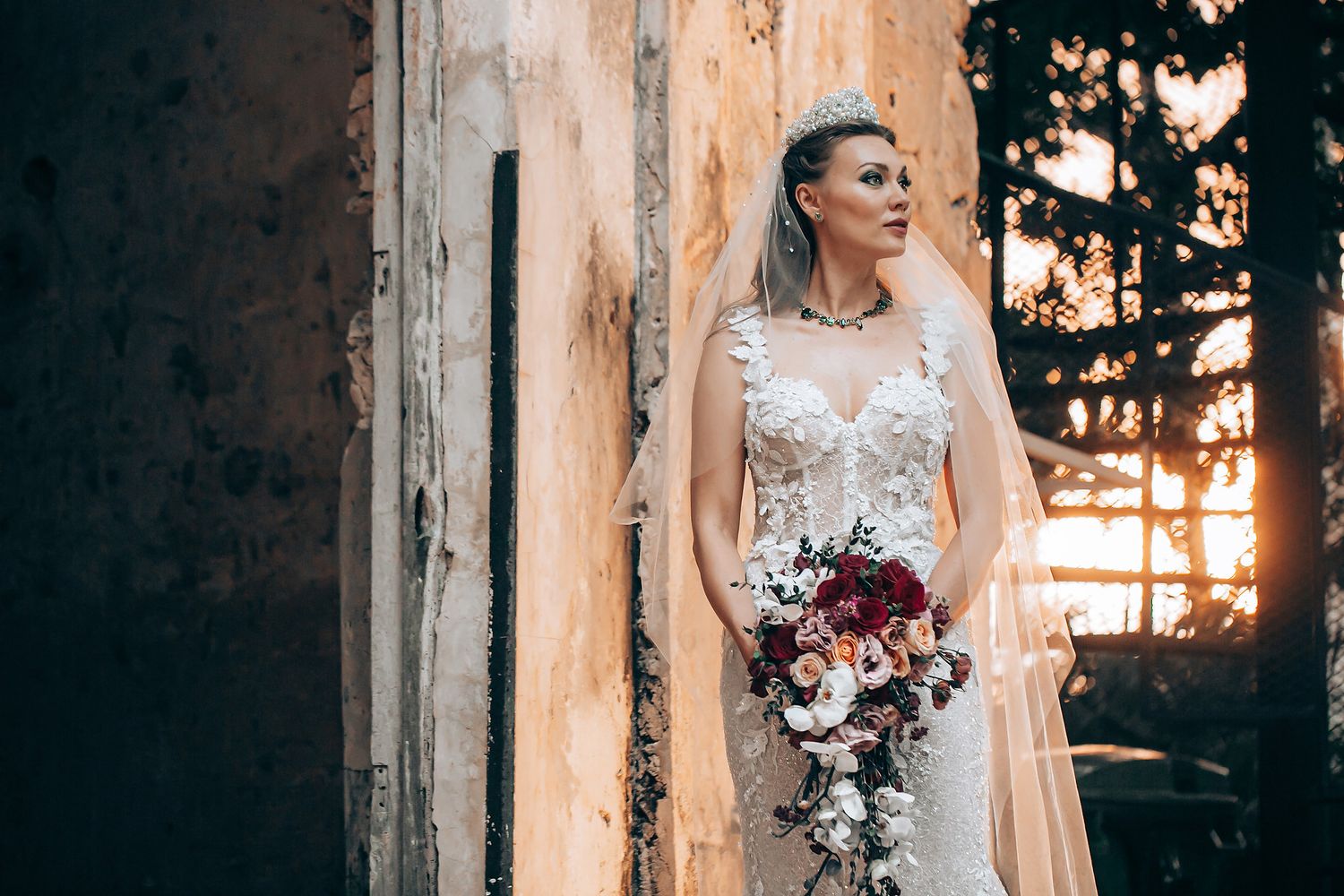 Bride in white lace wedding dress holds bouquet while posing against rustic stone wall at sunset.