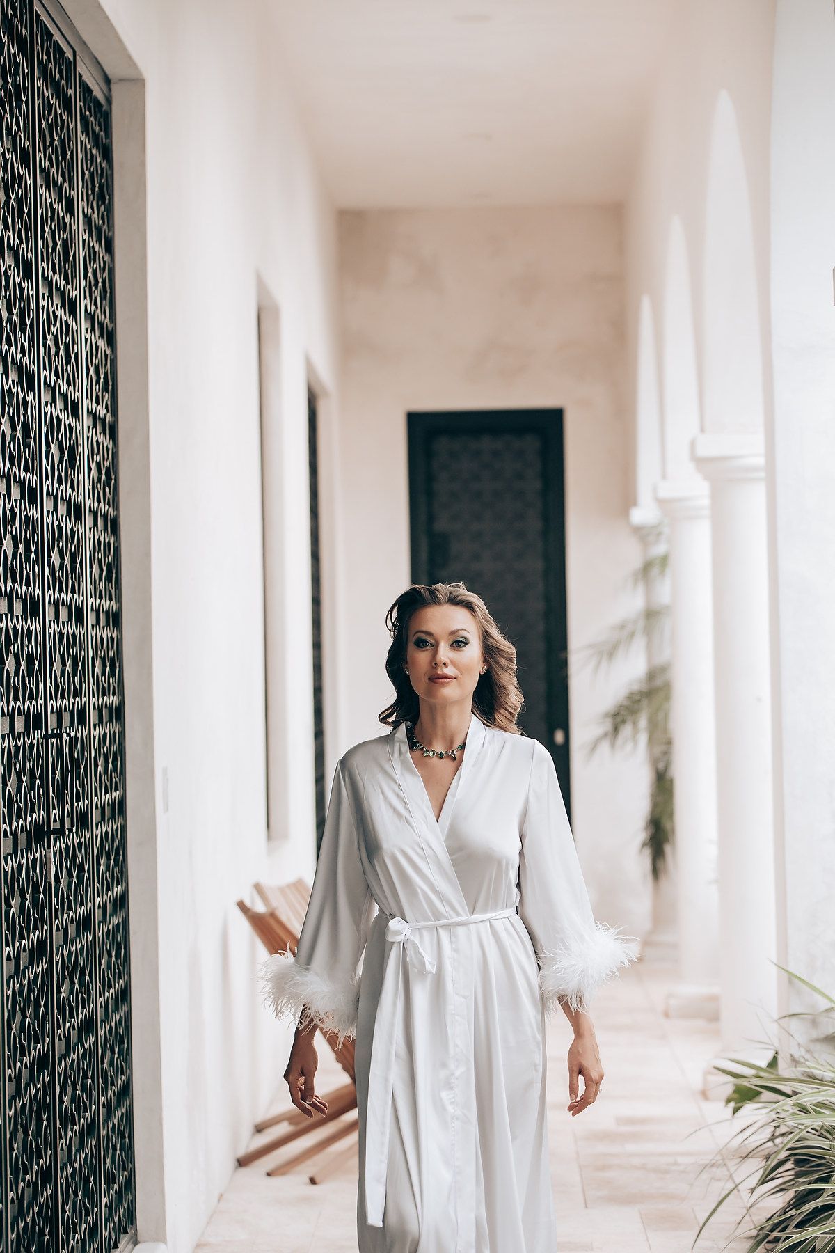 A person in a flowing white dress walks through an elegant white corridor with ornate iron railings and natural light.