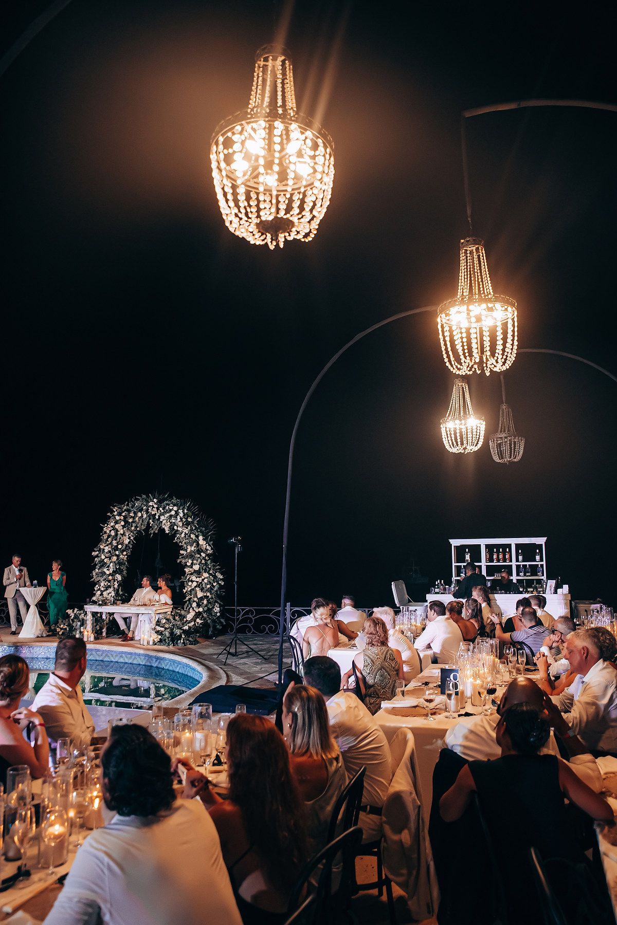 Elegant wedding reception dinner under crystal chandeliers with guests seated at long tables in dimly lit venue.