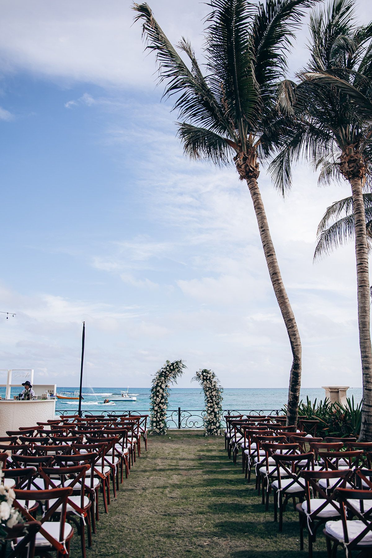 Beachfront wedding ceremony setup with wooden chairs and floral arch between palm trees overlooking the ocean.