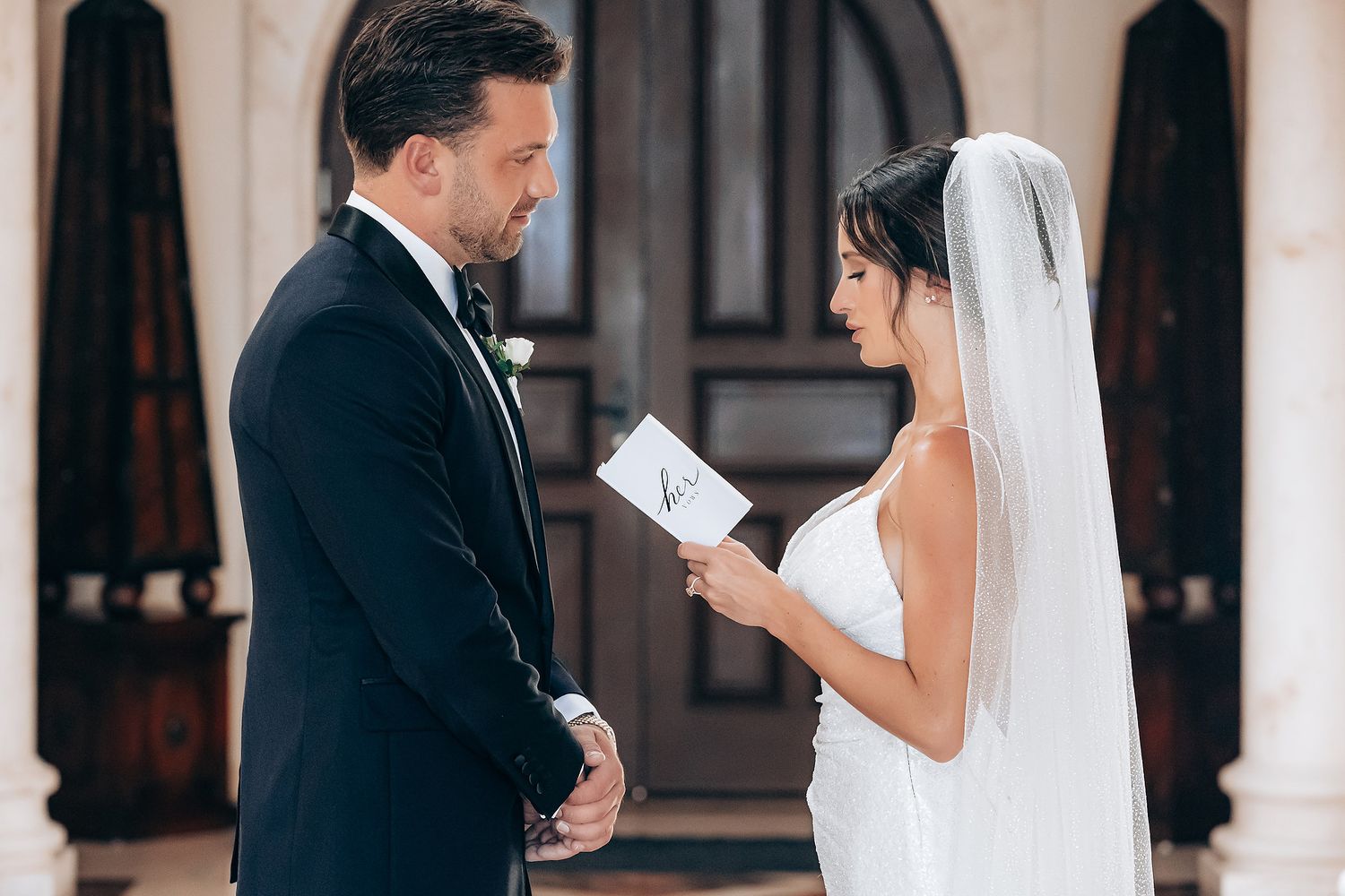 Bride and groom exchange vows in an elegant white columned corridor with dramatic lighting.