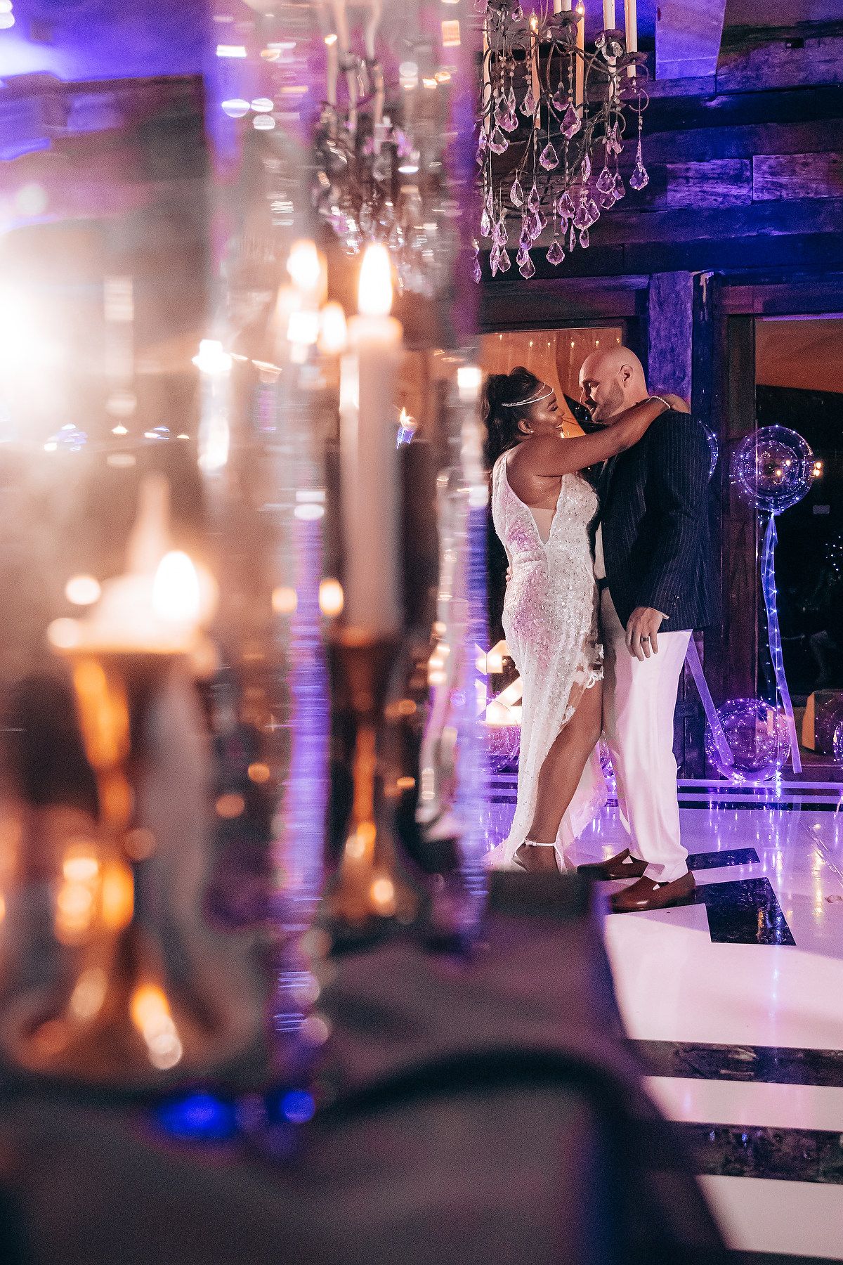 Elegant wedding reception scene with dancers on a white dance floor illuminated by purple lighting and crystal chandeliers.
